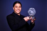 A woman in a black suit smiles and holds up a glass trophy shaped like a soccer ball.