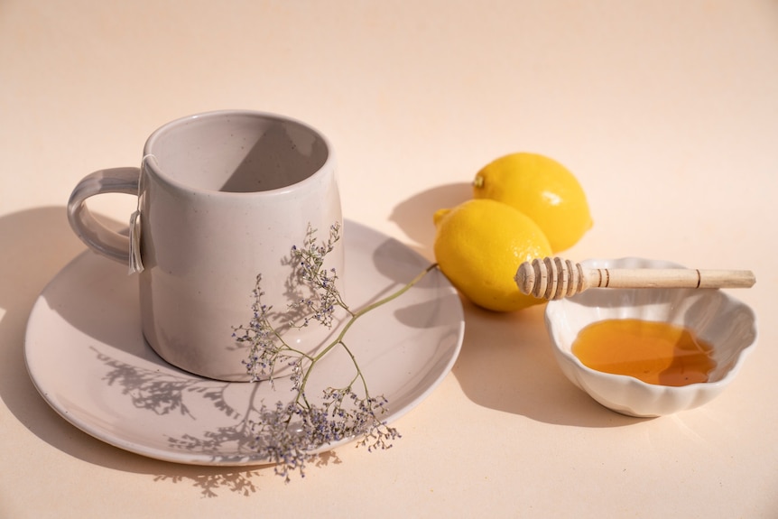 A tea cup and saucer placed next to two fresh lemons and a little dish of honey