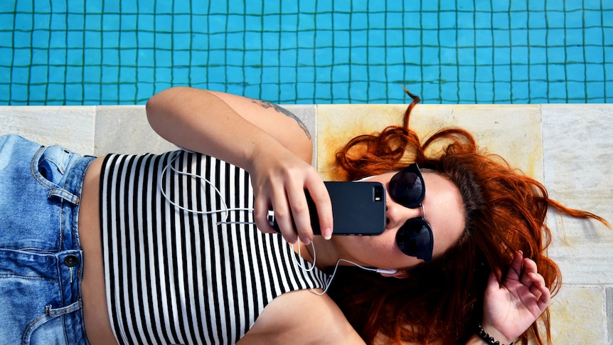 Next to just visible blue pool water, a woman with sunglasses and stripy top lays with phone held over her face.