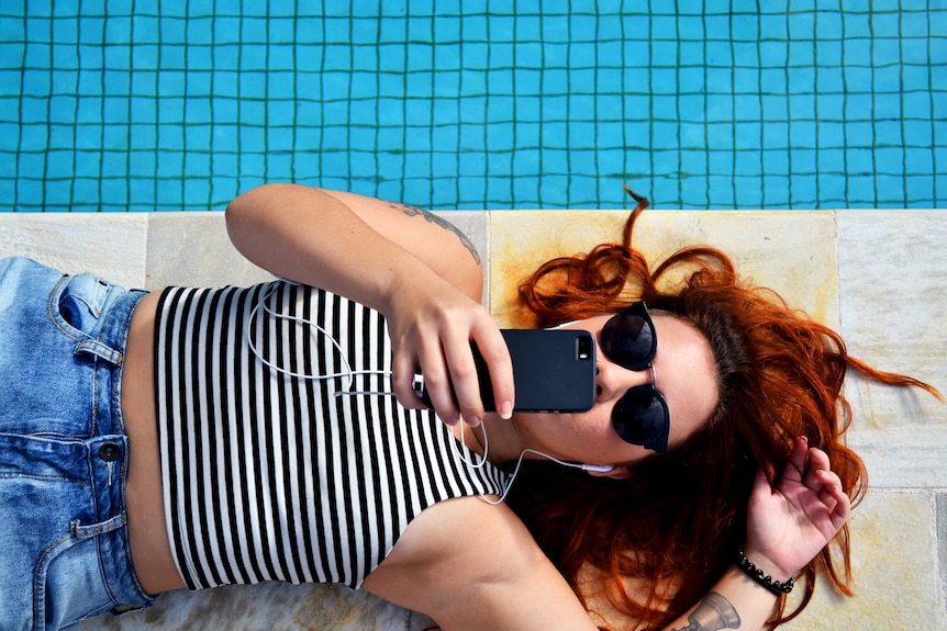 Next to just visible blue pool water, a woman with sunglasses and stripy top lays with phone held over her face.
