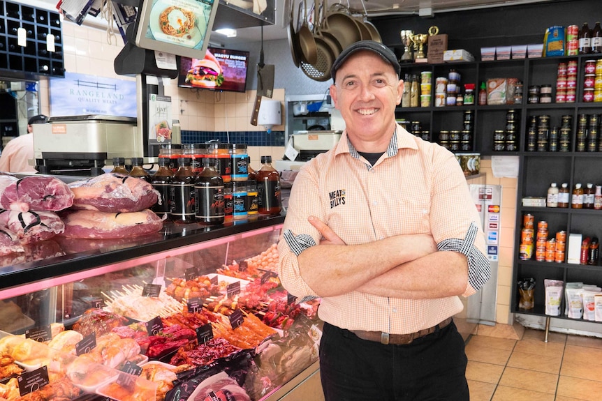 A man stands, armed folded and smiling, in front of a counter inside a butcher.
