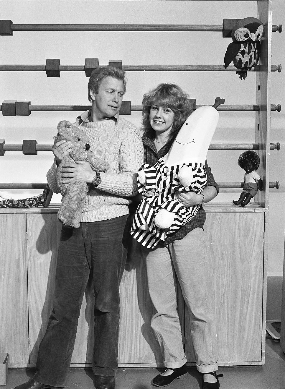 Black and white photos of John Hamblin and Noni Hazelhurst.  They are both holding soft toys on the Play School set.