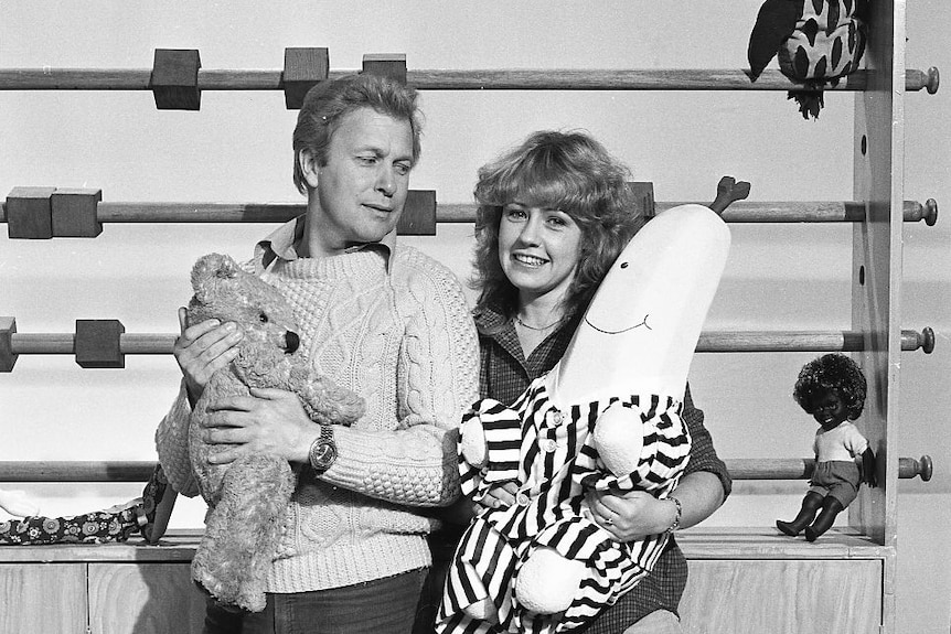 Black and white photos of John Hamblin and Noni Hazelhurst. They are both holding soft toys on the Play School set.