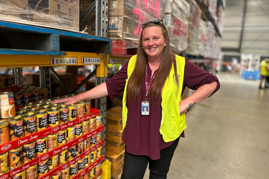 Woman in high vis vest smiles standing in front of shelves filled with tinned food.