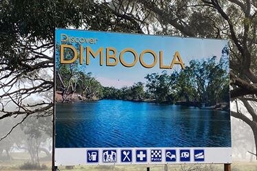 a blue sign that says dimboola with a picture of  lake. the sign is in front of scrub land with fog