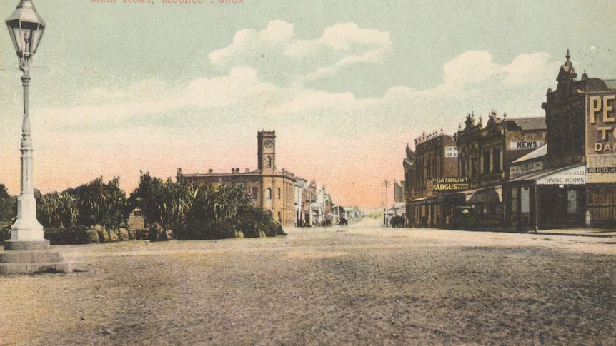 Panoramic streetscape from 1900, ornate lamppost beside a landscaped island in a wide, unsurfaced road, clock tower.
