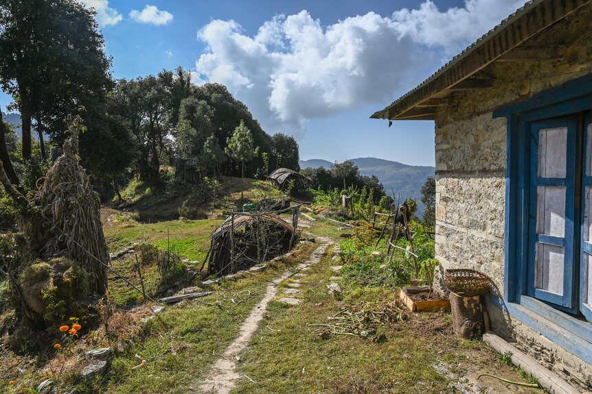 Farm life is visible next to a stone house with blue windows and mountains in the distance