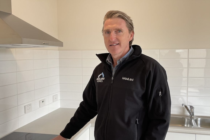 A slightly smiling middle-aged man, light hair, a black jacket with logo, stands in an empty kitchen with white tiles.