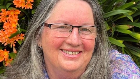 woman with long grey hair with lilac paisley scarf in front of bush with orange flowers