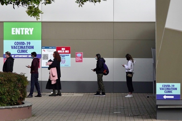 Personnes attendant à l'extérieur d'un bâtiment avec des panneaux indiquant ENTREE à la clinique de vaccination COVID-19