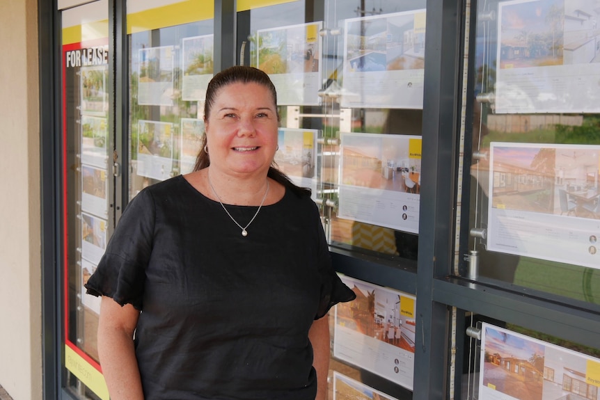 A middle-aged woman with long, dark hair stands outside a real estate agency.