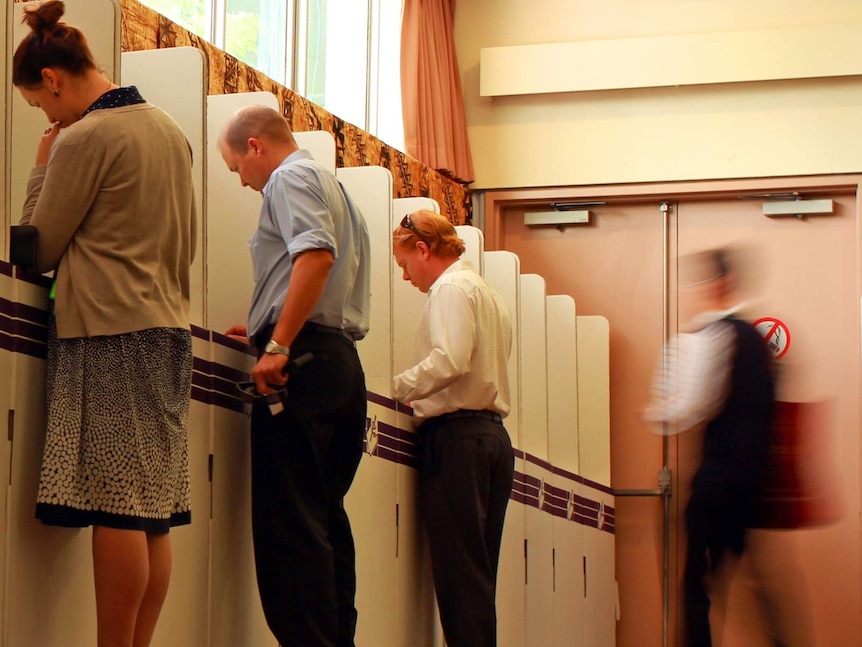 Three unidentified voters at a polling booth and a blurry walking figure heads to cast vote