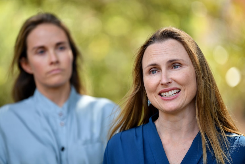 a young woman speaking to the media outdoors with another young woman standing behind her