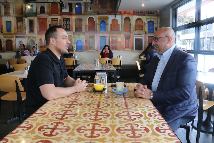 Two men in cafe having coffee, facing each other. Wallpaper in background shows different colourful doorways.