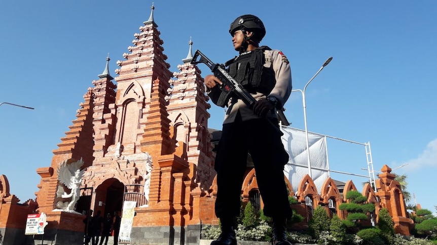 An Indonesian police officer stands with a high-powered weapon in front of a chuch.