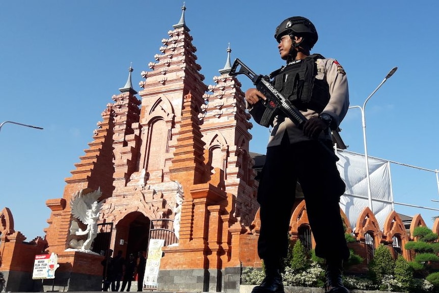 An Indonesian police officer stands with a high-powered weapon in front of a chuch.