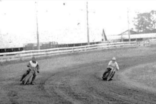 A black and white image of two men on motorbikes going around a dirt track