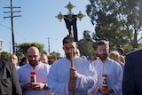 Three Maronite altar servers dressed in white robes bow their heads. One holds gold cross.
