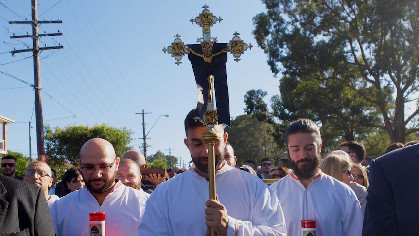 Three Maronite altar servers dressed in white robes bow their heads. One holds gold cross.