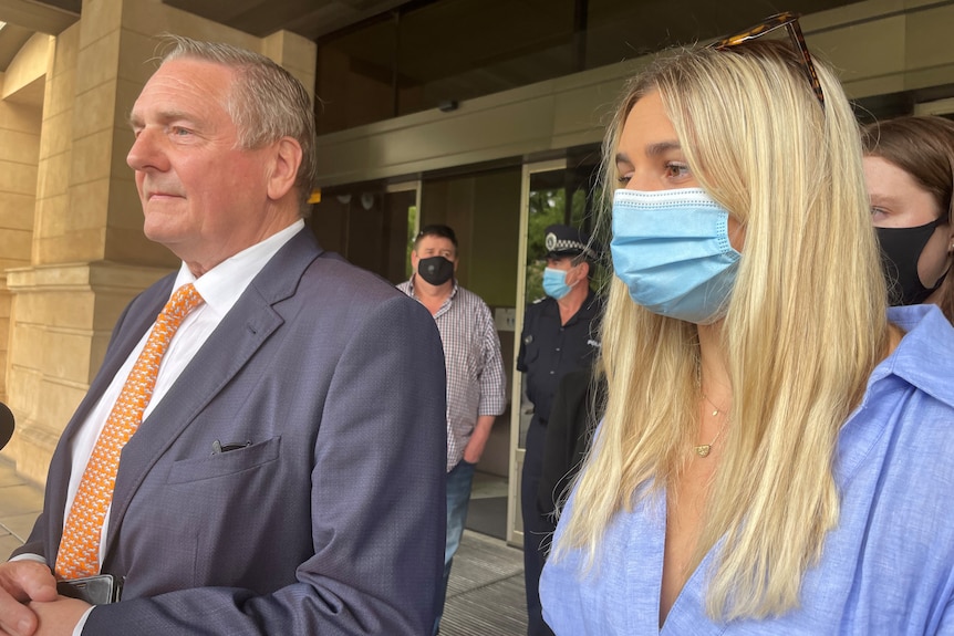 A man in a suit stands next to a woman with long blonde hair wearing a face mask