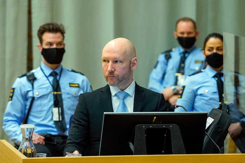 A bald man wearing a suit and blue tie sits behind a computer flanked by security guards wearing face masks.