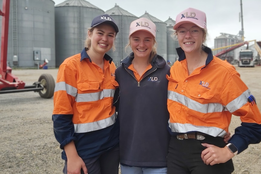 Three smiling young women stand in front of a row of large grain silos in high vis clothes.