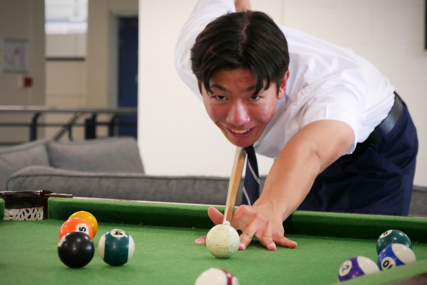 A young man in formal school uniform bends over a pool table and aims a pool cue at a white ball.