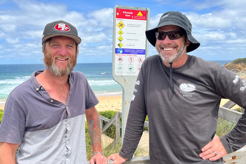 Two smiling, bearded men in hats standing in front of a beach.