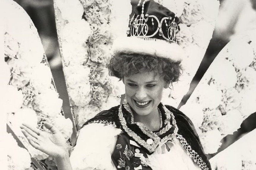 A smiling woman sits on a parade float chair wearing a crown and cloak, waving.