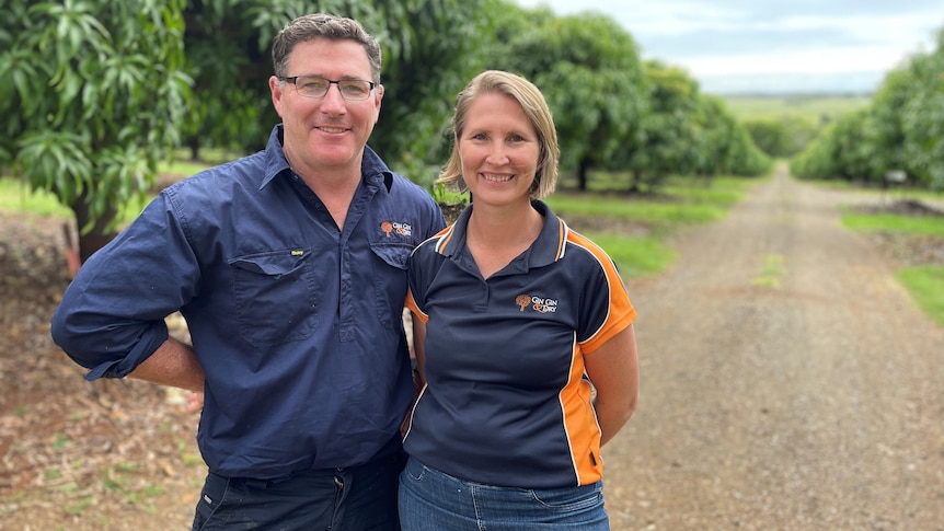 Photo of a man and a woman smiling in front of an orchard.