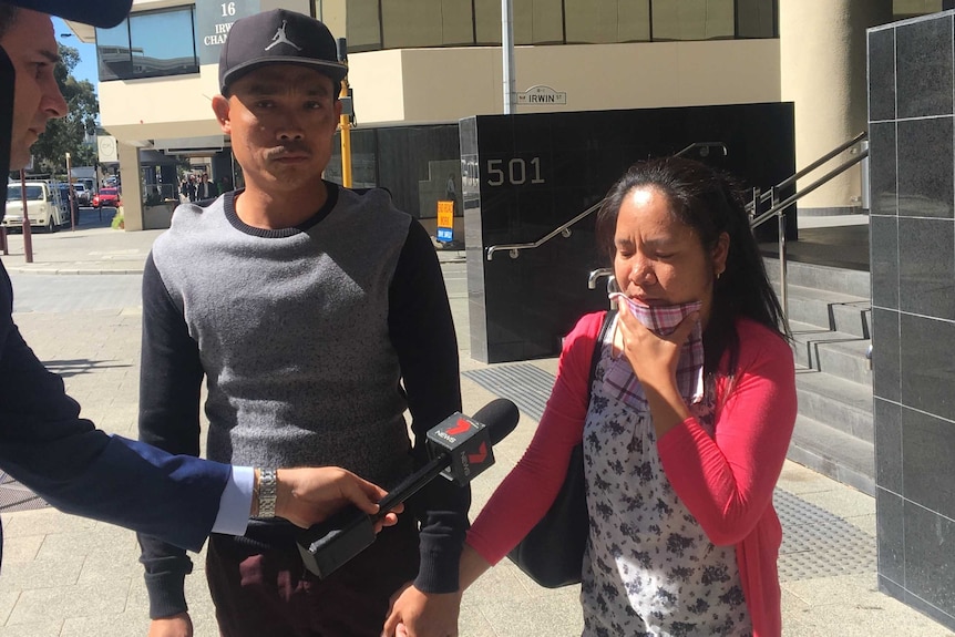 A woman in a red top holds the hand of a man in a black cap and top outside court while being questioned by media.