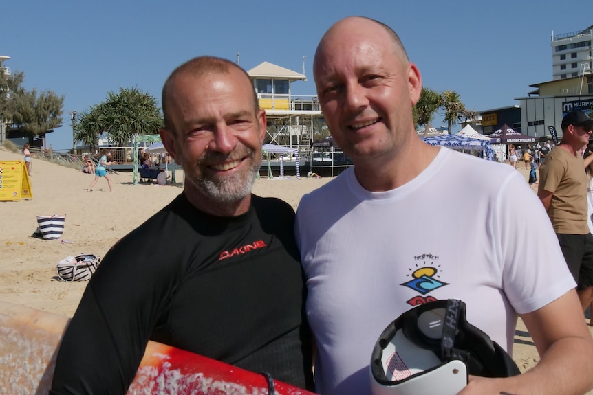 Two men in rash vests smile while holding surfboards on a beach.