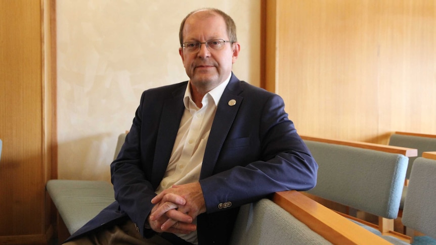 A man in a suit sits in a funeral home, looking at the camera.