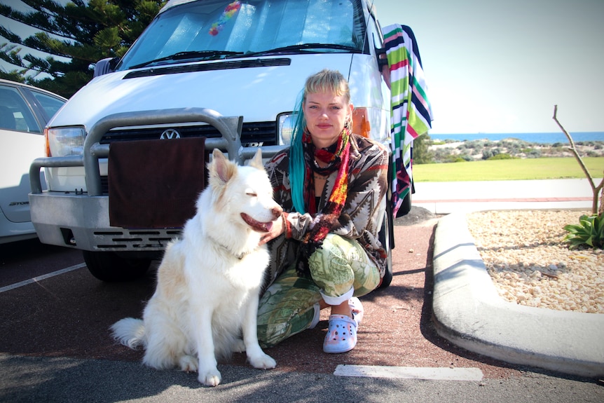 A blonde lady kneels down beside a dog in front of a van