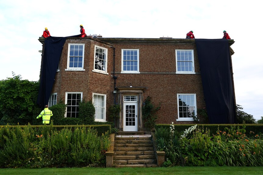 A two storey house with four people wearing orange on the roof covering the building with large black sheets of fabric