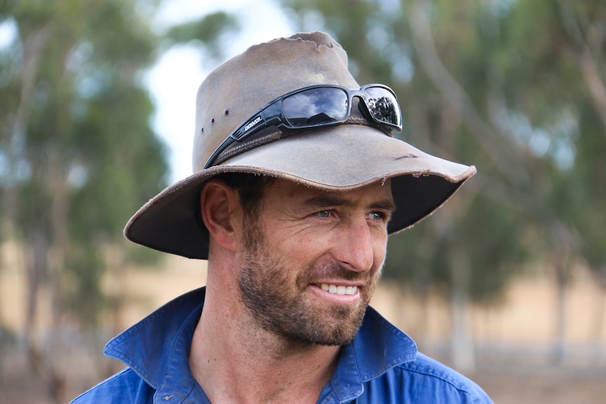 Man in a dark blue collared shirt wears a worn brimmed hat with sunglasses on top. He looks to the side, smiling.