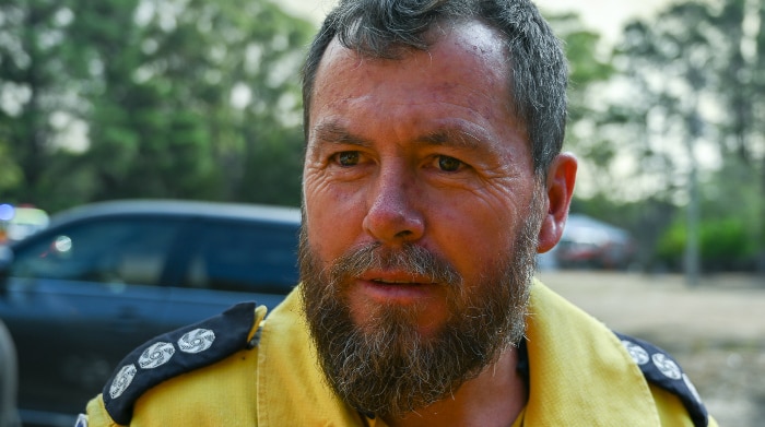 RFS firefighter Andrew Beville stands in front of bushland and a fire truck.