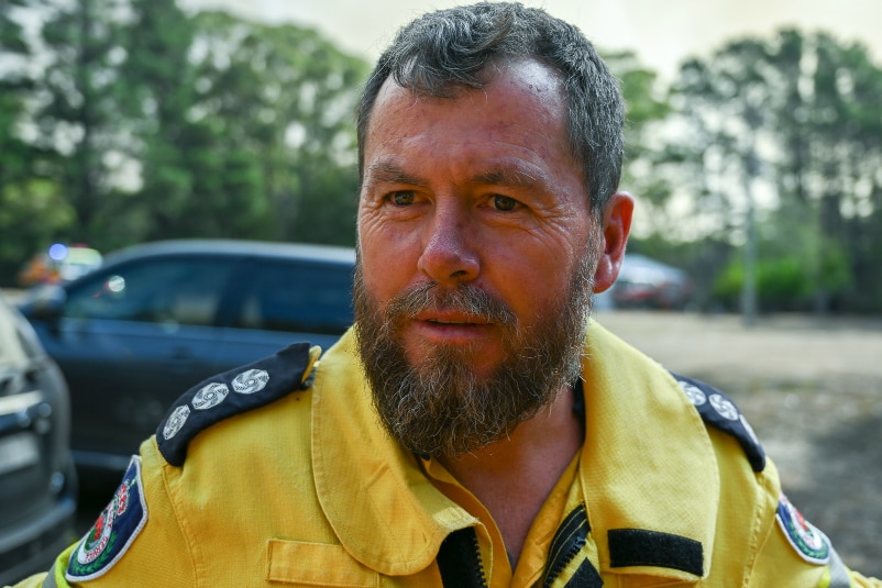 RFS firefighter Andrew Beville stands in front of bushland and a fire truck.