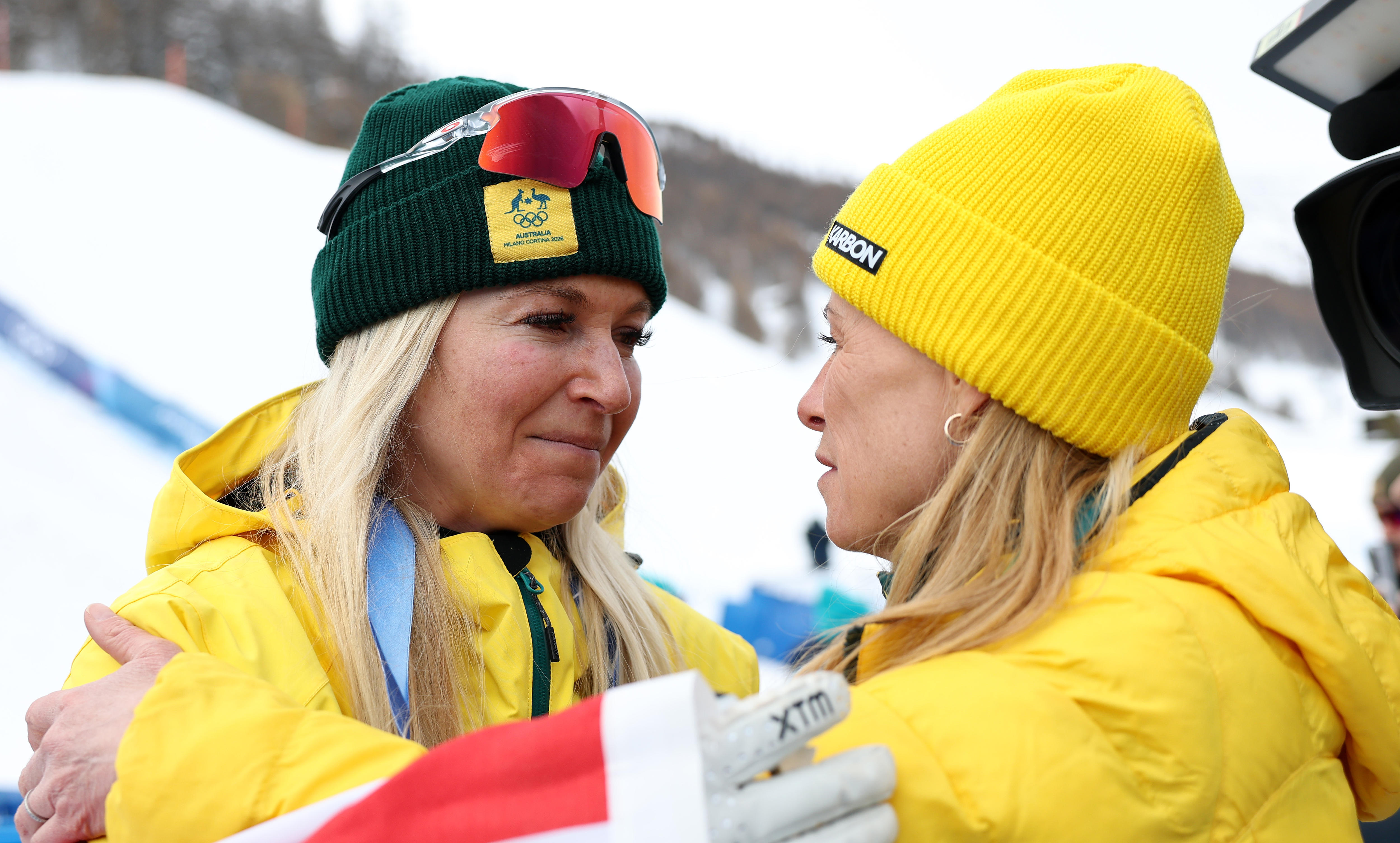 Two blonde women in beanies and parkas share a moment on a snowfield.