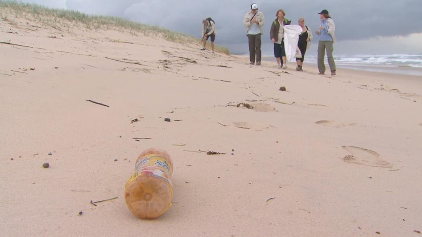 People stand on beach, discarded drink bottle on sand