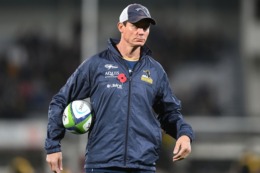 The Brumbies' 2017 coach Stephen Larkham stands with a ball under his left arm.
