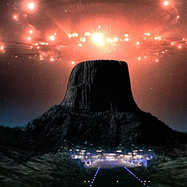 An image of a mountain-top with a flat surface where a huge red-lit alien spaceship is landing.