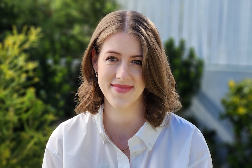 A women with shoulder length wavy brown hair and a white shirt, looks at the camera, greenery behind. 