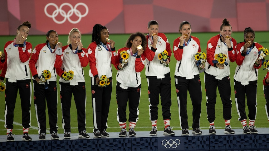 A female soccer team stands on a podium wearing tracksuit pants, holding medals and flowers after winning a tournament