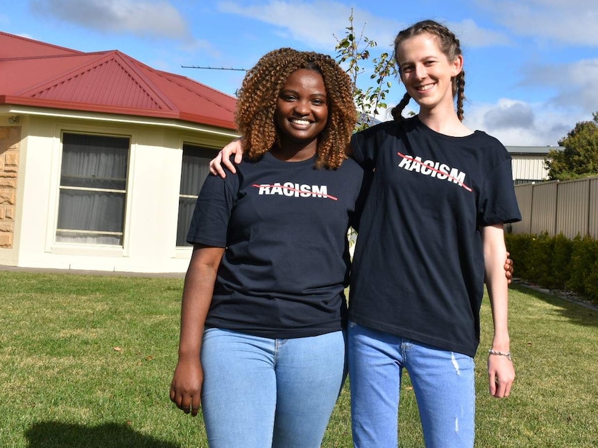 Chanceline Kakule, left, puts her arm around Lily Coote, right, as they stand in front of a house.