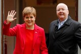 Nicola Sturgeon, wearing red, waves at the media alongside her husband Peter Murrell.