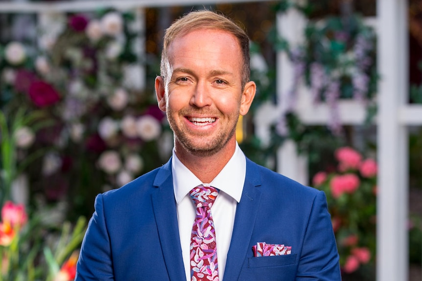 Promotional photo of Jess Glasgow smiling in a suit on The Bachelorette 2019 television program.