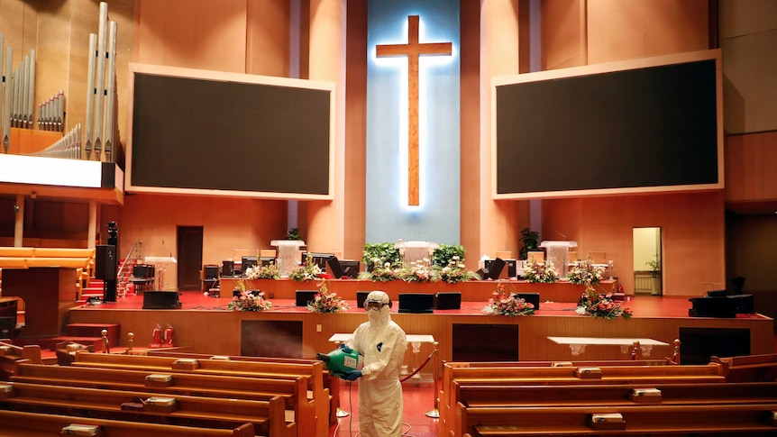 A man dressed in a white safety suit and wearing safety goggles and a mask stands holding a spray in a church.