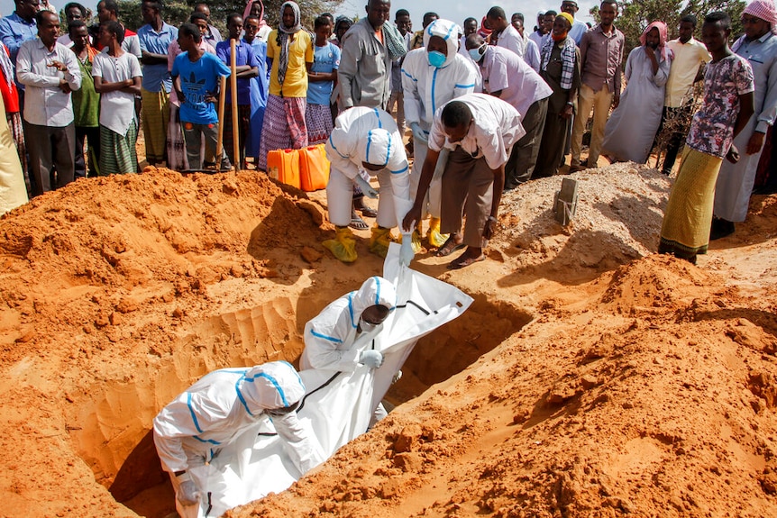 Four people in protective suits, and one in regular clothes, help shift a body bag into a grave. A crowd watches from behind.