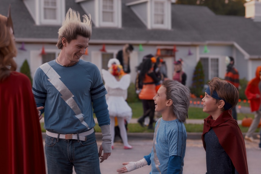 All dressed in superhero costumes, a woman faces away while a 30-something man smiles down at two children on Halloween.